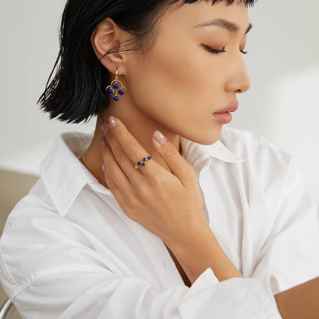 A woman wearing a white shirt and earrings with blue stones is shown in profile, her hand resting on her chin.